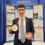 Young man in a suit holding a first-place medal and a colorful award in front of a M.A.N.T.I.S research poster booth.