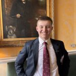 Smiling young man in a suit posing in front of a large framed portrait in a yellow-walled room with patterned wallpaper and a dog painting in the frame's lower left corner.