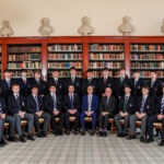 Group of students in blazers posing in a library with tall bookshelves and busts on display above the shelves.