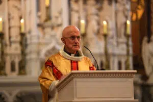 Elderly priest in gold vestments delivering a sermon at a white pulpit inside a church.
