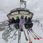 Group of skiers on a chairlift above a snowy mountain landscape, wearing helmets and colorful jackets.