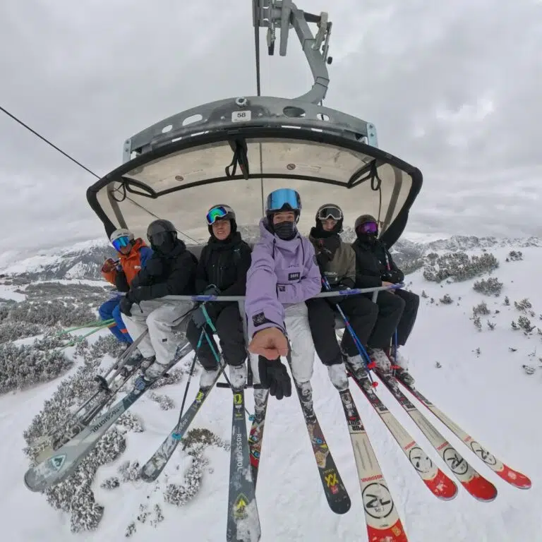 Group of skiers on a chairlift above a snowy mountain landscape, wearing helmets and colorful jackets.